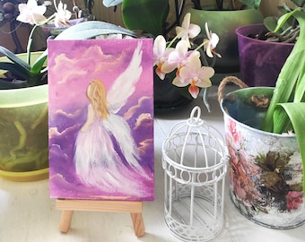 May include: A small painting on a wooden easel depicts an angel with white wings flying through a purple and pink cloudy sky.