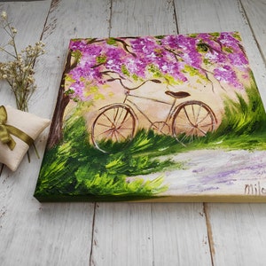 May include: A painting of a bicycle parked under a tree with purple flowers. The bicycle is white with brown wheels and a brown seat. The background is a green field with a path leading to the bicycle.