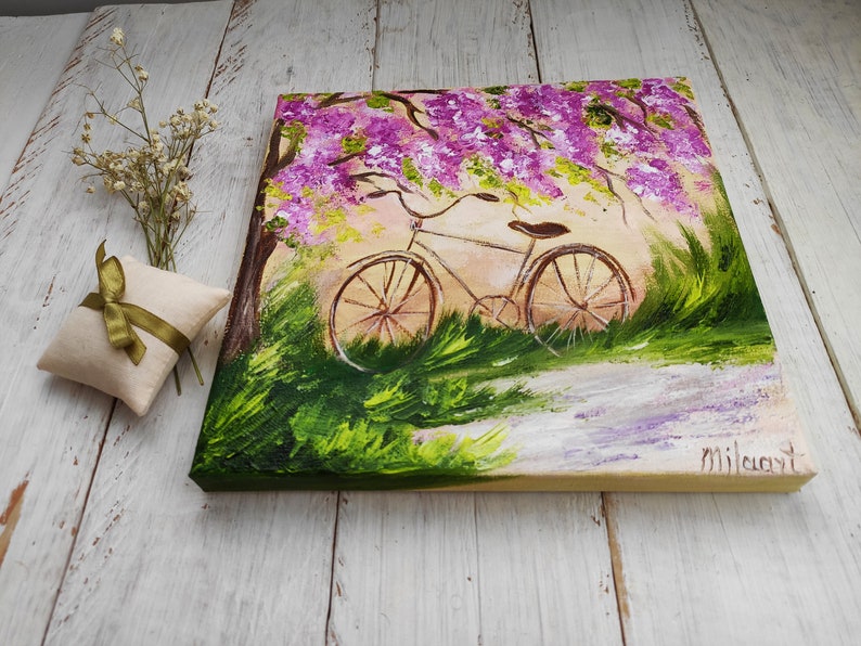 May include: A painting of a bicycle parked under a tree with purple flowers. The bicycle is white with brown wheels and a brown seat. The background is a green field with a path leading to the bicycle.
