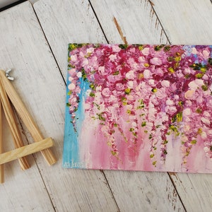 K&ouml;nnte beinhalten: Ein kleines Gem&auml;lde auf Leinwand mit einem Holzstaffelei. Das Gem&auml;lde zeigt eine Ansammlung von rosa Bl&uuml;ten mit gr&uuml;nen Bl&auml;ttern, die vom oberen Rand der Leinwand herabh&auml;ngen. Der Hintergrund ist ein hellblauer und rosafarbener Farbverlauf.