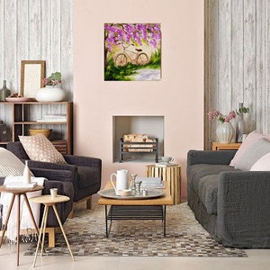 May include: A painting of a bicycle parked under a tree with pink flowers. The bicycle is white with brown tires and a brown seat. The tree is in the background and the flowers are in bloom. The painting is in a living room setting with a fireplace and two couches.