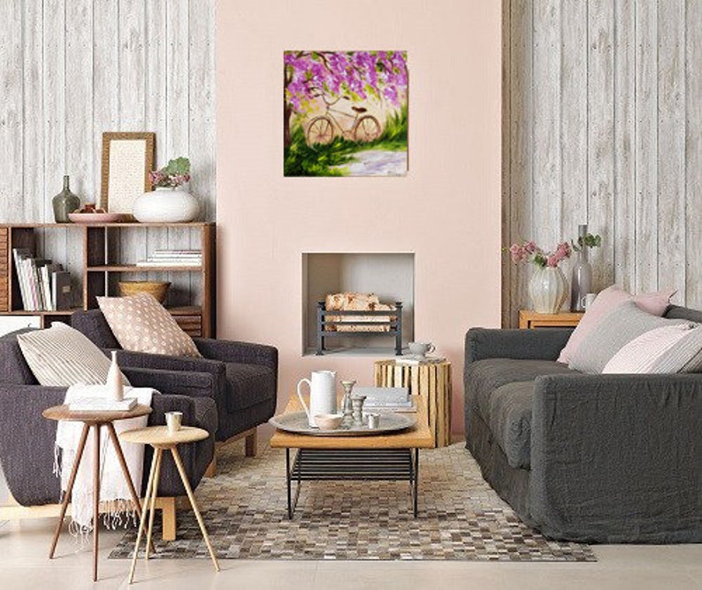 May include: A painting of a bicycle parked under a tree with pink flowers. The bicycle is white with brown tires and a brown seat. The tree is in the background and the flowers are in bloom. The painting is in a living room setting with a fireplace and two couches.