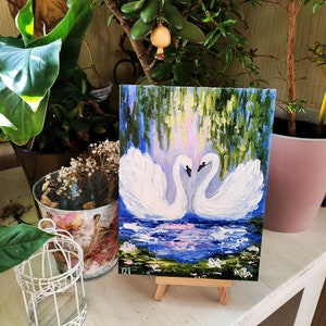 May include: A painting of two white swans with their necks intertwined, set against a blue and green background with water lilies. The painting is on a small easel.