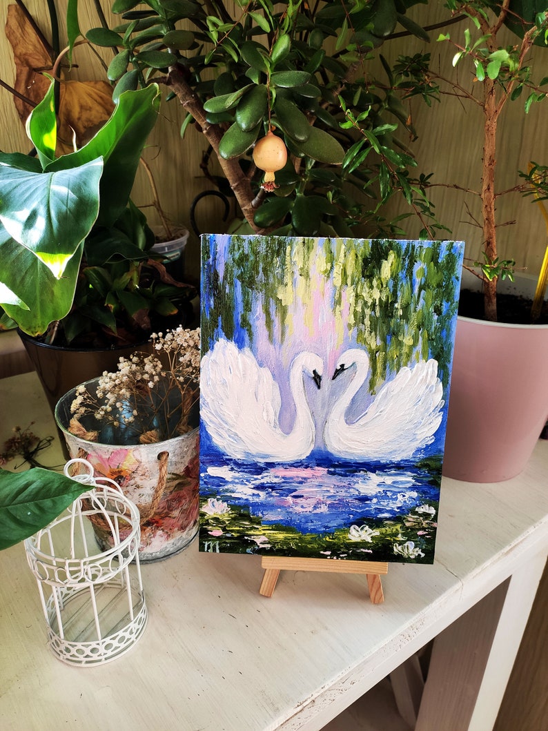 May include: A painting of two white swans with their necks intertwined, set against a blue and green background with water lilies. The painting is on a small easel.