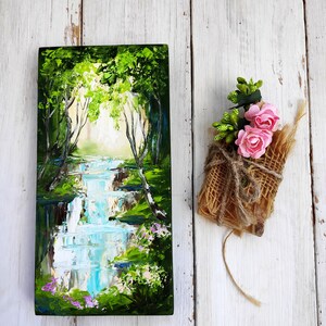 Op de afbeelding: Een schilderij van een boslandschap met een waterval en groene bomen. Het schilderij is op een houten paneel en is zwart ingelijst. Er is een klein boeket roze bloemen verpakt in jute naast het schilderij.