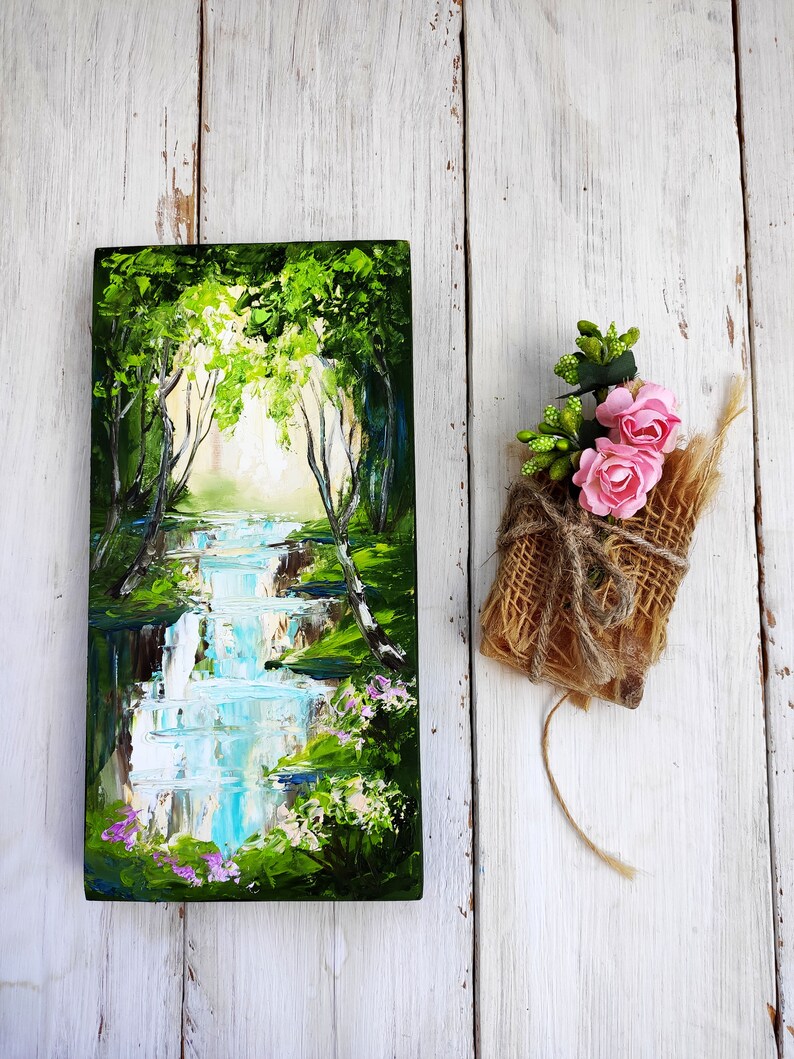 Op de afbeelding: Een schilderij van een boslandschap met een waterval en groene bomen. Het schilderij is op een houten paneel en is zwart ingelijst. Er is een klein boeket roze bloemen verpakt in jute naast het schilderij.