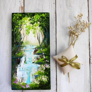 Op de afbeelding: Een klein schilderij op een houten paneel toont een weelderige groene bos sc&egrave;ne met een waterval die er doorheen stroomt. Het schilderij heeft een lichtblauwe lucht en een heldere witte zon die door de bomen schijnt.