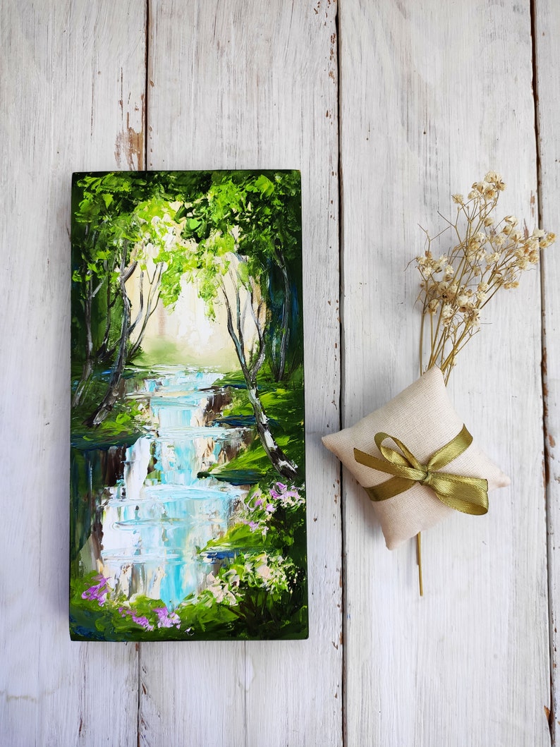 Op de afbeelding: Een klein schilderij op een houten paneel toont een weelderige groene bos sc&egrave;ne met een waterval die er doorheen stroomt. Het schilderij heeft een lichtblauwe lucht en een heldere witte zon die door de bomen schijnt.