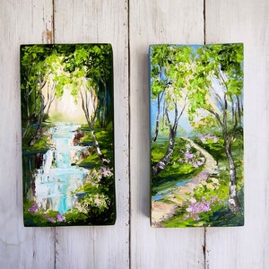 Op de afbeelding: Twee kleine schilderijen op een witte houten achtergrond. Het schilderij aan de linkerkant toont een waterval in een groen bos. Het schilderij aan de rechterkant toont een pad door een bos met berkenbomen en roze bloemen.