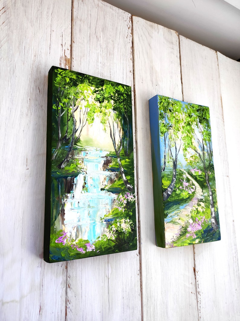 Op de afbeelding: Twee ingelijste schilderijen die natuursc&egrave;nes uitbeelden. Het schilderij aan de linkerkant toont een waterval in een groen bos. Het schilderij aan de rechterkant toont een pad door een berkenbos.