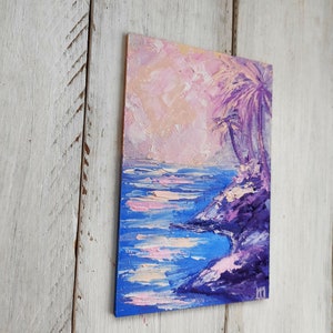 Könnte beinhalten: Ein kleines Gemälde, das eine violette und blaue Meereslandschaft mit Palmen im Hintergrund zeigt. Der Himmel ist in einem zarten Rosa und Orange gehalten.