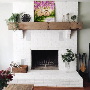 May include: White brick fireplace with a wooden mantel and a painting of a bicycle under a blooming tree above the fireplace.