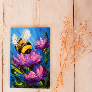 May include: A painting of a bumblebee flying near purple flowers with yellow centers. The background is a blue sky. The painting is on a wooden surface.