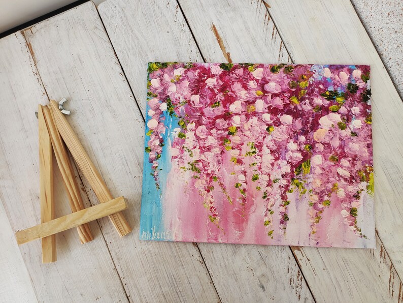 K&ouml;nnte beinhalten: Ein kleines Gem&auml;lde auf Leinwand mit einem Holzstaffelei. Das Gem&auml;lde zeigt eine Ansammlung von rosa Bl&uuml;ten mit gr&uuml;nen Bl&auml;ttern, die vom oberen Rand der Leinwand herabh&auml;ngen. Der Hintergrund ist ein hellblauer und rosafarbener Farbverlauf.