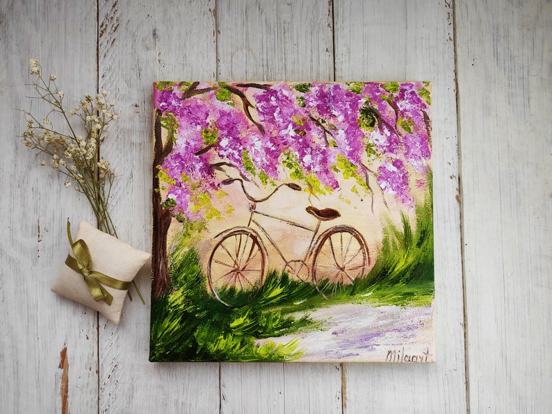 May include: A painting of a bicycle parked under a tree with pink flowers. The bicycle is brown with a white seat and has a basket on the handlebars. The background is a light blue sky with white clouds.