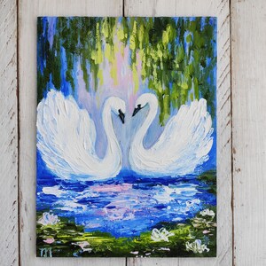 May include: A painting of two white swans with their necks intertwined, facing each other. The swans are in a blue pond with green lily pads and white flowers. The background is a blue sky with green trees.