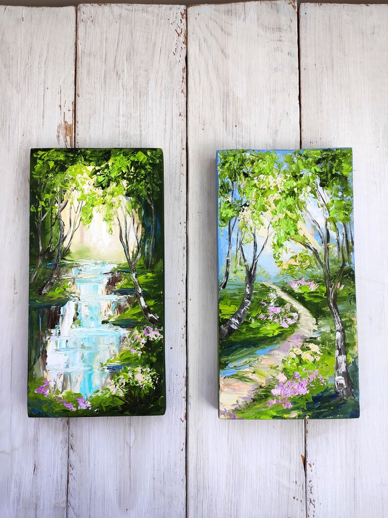 Op de afbeelding: Twee kleine schilderijen op canvas, die elk een andere sc&egrave;ne in een bos voorstellen. Het schilderij links toont een waterval in een groen bos met een blauwe lucht erboven. Het schilderij rechts toont een pad dat door een groen bos slingert met een blauwe lucht erboven.