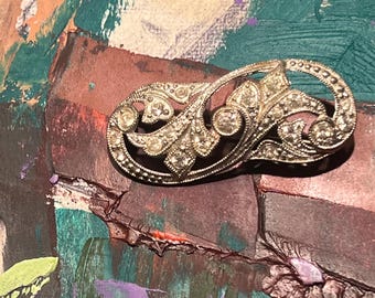 May include: A vintage silver-toned brooch with a floral design, encrusted with clear rhinestones. The brooch has an ornate, curved shape with detailed embellishments. The background features a colorful abstract painting with shades of green, purple, and brown.