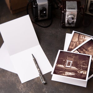 May include: A still-life photograph featuring vintage cameras, blank greeting cards, and sepia-toned prints. A patterned pen rests near the open cards. The prints depict antique cameras and other vintage objects. The scene is set on a dark, textured surface.