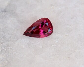 May include: A pear-shaped, faceted gemstone with a deep pink hue. The gemstone is set against a white background.