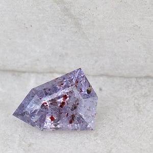 以下が含まれることがあります： 赤色のインクルージョンが入った、ファセットカットされた薄紫色の宝石。この宝石は、複数のファセットを持つユニークな幾何学的な形状をしています。石は白いテクスチャード加工の背景に配置され、その色と透明度を際立たせています。この宝石は、おそらくジュエリーに使用されています。