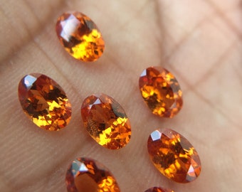 May include: Seven oval-shaped orange gemstones with a reddish hue. The gemstones are arranged in a semi-circle on a light brown surface.