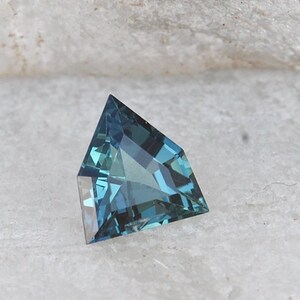 May include: A kite-shaped gemstone, displaying a range of teal and blue hues. The facets within the gem create a captivating interplay of light. The stone is set against a pale background, accentuating its colour and cut, typical of a precious stone.