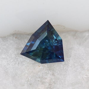 Pode incluir: Uma gema azul escura com forma triangular facetada. A gema está colocada sobre um fundo branco.