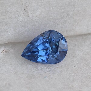 Puede incluir: Una gema facetada en forma de pera, de un tono azul vibrante. La superficie de la gema refleja la luz, mostrando sus intrincados cortes y ángulos. La piedra se encuentra sobre un fondo neutro y texturizado.