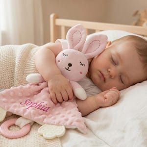 Könnte beinhalten: Ein schlafendes Baby kuschelt mit einem rosa Hasen-Kuscheltuch. Das Kuscheltuch hat eine strukturierte, gepunktete Oberfläche und den Namen "Sophia" aufgestickt. Der Hase hat rosa Ohren und ein süßes Gesicht.