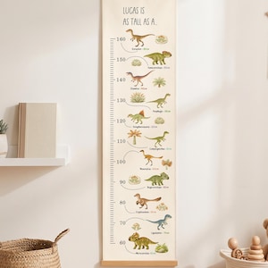 Puede incluir: Un medidor de altura vertical de lienzo con rieles de madera y un cordón para colgar. El medidor presenta el texto "LUCAS IS AS TALL AS A..." y una escala métrica de 60 a 160 cm, ilustrada con varias especies de dinosaurios coloridos y sus nombres.
