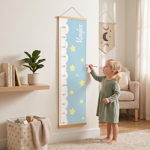 May include: A vertical wall-mounted growth chart featuring a light blue background with yellow stars, a white cloud-shaped border, and black measurement markings from 60 to 160 centimetres. The name Kaylee is printed in white script at the top.
