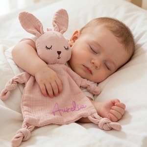 Puede incluir: Un bebé dormido abraza una mantita de seguridad con forma de conejito rosa. El conejito tiene orejas largas y ojos cerrados. La mantita está personalizada con el nombre "Aurelia" en cursiva. El bebé está sobre una almohada blanca.