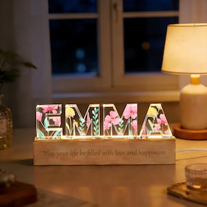 Peut inclure: Un panneau décoratif lumineux épelant "EMMA" avec des lettres en acrylique transparent remplies de motifs floraux roses et jaunes. Le panneau repose sur une base en bois avec le texte gravé "May your life be filled with love and happiness."