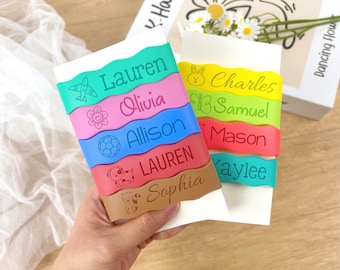 May include: A selection of colourful name bands, each with a different name and icon. The bands are in shades of green, pink, blue, red, and brown. Each band features a unique name and icon, such as a flower, airplane, soccer ball, dog, and cat.