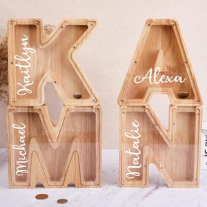 Può includere: Salvadanai a forma di lettera in legno. Le lettere sono K, M, A e N, con nomi come "Kaitlyn", "Michael", "Alexa" e "Natalie" in scrittura bianca. Ogni salvadanaio ha un pannello frontale trasparente e una fessura per le monete. Il legno è di colore marrone chiaro.
