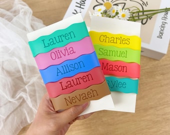 Könnte beinhalten: Zwei personalisierte Namensschilder mit farbenfrohen, wellenförmigen Bändern. Ein Schild hat Namen wie "Lauren" und "Olivia" in Türkis, Rosa, Blau, Rot und Braun. Das andere Schild hat Namen wie "Charles" und "Samuel" in Gelb, Grün, Rot und Türkis.