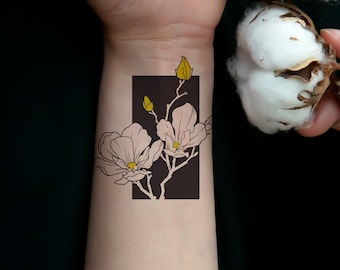 May include: A temporary tattoo featuring a floral design of light pink magnolia blossoms with yellow centers and buds, set against a dark brown rectangular background. The tattoo is applied to the wrist.