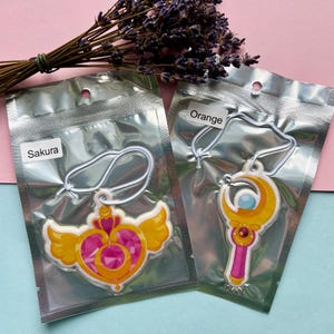 May include: Two car air fresheners in clear plastic packaging. The left air freshener is shaped like a heart with wings and has a pink gem in the centre. It is labelled "Sakura". The right air freshener is shaped like a wand with a crescent moon and a blue gem. It is labelled "Orange".