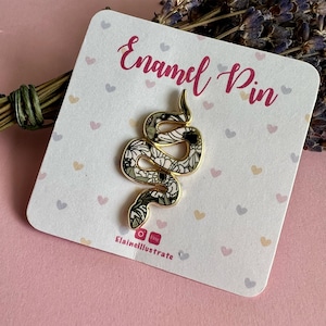 May include: A gold-outlined enamel snake pin with a black, white, and green floral design, displayed on a white card. The card features the words "Enamel Pin" in pink script and small heart shapes. The pin is a decorative accessory.