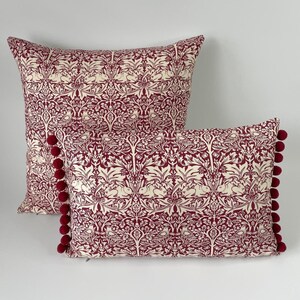 Può includere: Due cuscini decorativi con sfondo bordeaux e un motivo floreale bianco. I cuscini hanno un bordo con pompon bordeaux.