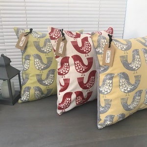 May include: Three decorative throw pillows with a bird pattern. The pillows are in yellow, red, and gray. The pillows are on a gray surface.