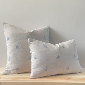 May include: Two beige cushions featuring a repeating pattern of blue sailboats. The cushions are rectangular and have a textured appearance. They are placed on a light-coloured wooden surface.