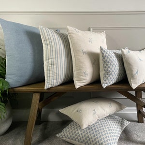 Op de afbeelding: Een verzameling decoratieve kussens in verschillende patronen en kleuren, waaronder blauw, wit en beige. De kussens zijn gerangschikt op een houten bank.