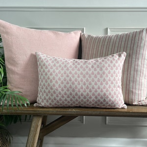 May include: Three decorative throw cushions on a wooden bench. The cushions are all pink and feature different patterns. The cushion on the left is a solid pink, the middle cushion has a small floral pattern, and the cushion on the right has a striped pattern.