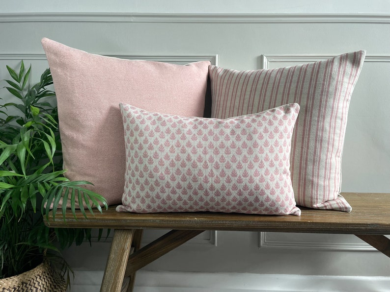 May include: Three decorative throw cushions on a wooden bench. The cushions are all pink and feature different patterns. The cushion on the left is a solid pink, the middle cushion has a small floral pattern, and the cushion on the right has a striped pattern.