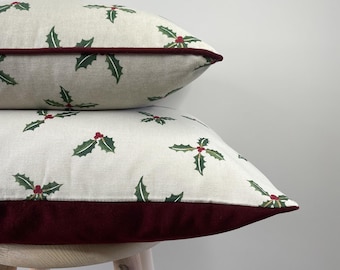 May include: Two rectangular throw pillows stacked on top of each other. The pillows feature a light beige fabric with a repeating pattern of green holly leaves and red berries. Each pillow is finished with a deep burgundy velvet piping trim.