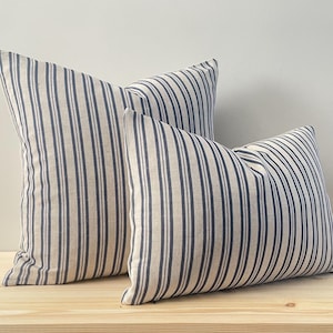 May include: Two decorative throw pillows with a striped pattern in shades of beige and navy blue. The pillows are made of a soft, textured fabric.