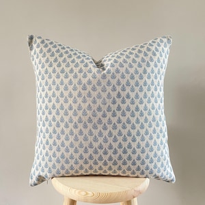 Op de afbeelding: Een vierkant decoratief kussen met een herhalend patroon van blauwe bladvormen op een crèmekleurige achtergrond. Het kussen staat op een kleine houten kruk. Het kussen heeft een zachte, getextureerde uitstraling.