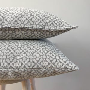 May include: Two decorative cushions with a grey and white geometric design, stacked on a light-coloured wooden stool. The cushions feature a repeating pattern of stylized floral shapes. The neutral colour scheme would suit a variety of interior styles.